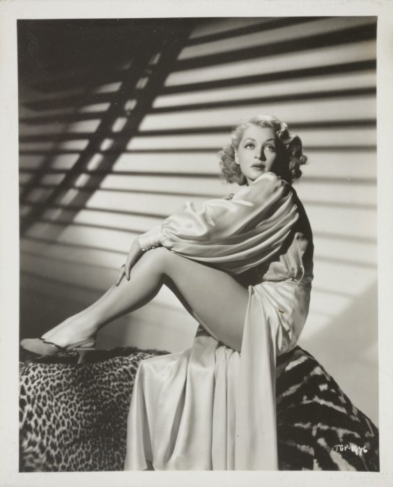 Promotional image of Lilli Palmer sitting on an animal print sofa in a long silk gown